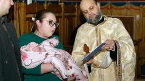 A woman holding a baby in a pink snowsuit while a priest reads